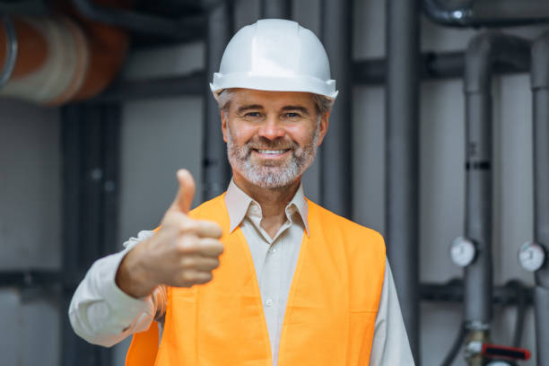 istock senior gray haired Smiling satisfied worker engineer in working clothes, with helmet and hardhat standing in factory and showing okay sign. thumb up in boiler room 1807903555