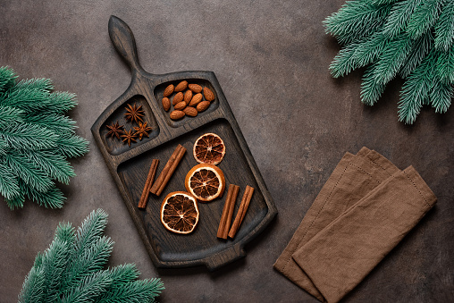 Christmas spices on a wooden cutting board decorated with decorative spruce branches, dark brown background. Top view, flat lay.