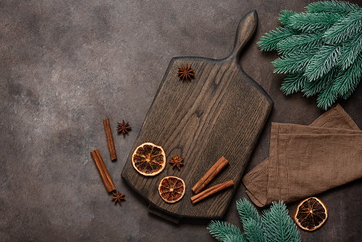 Culinary background with Christmas spices. Wooden cutting board, dry orange slices, cinnamon sticks, star anise and decorative fir branches on a dark brown background. Top view, flat lay.