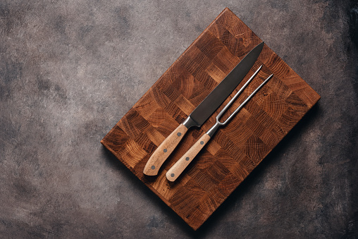 Oak cutting board with a knife and fork for meat on a dark rustic background. Top view, flat lay.
