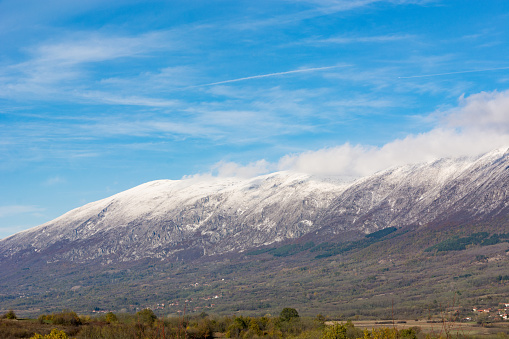 Witness the awe-inspiring grandeur of nature's landscape with this striking view. Towering tall and dry, the mountain dons its crown of snow, standing as an emblem of rugged beauty against the backdrop of a clear, sunny, and chilly autumn sky. The serene canvas, adorned with white clouds, complements the majestic mountain's silhouette. This image encapsulates the stark contrast between the snow-clad peak and the crisp, sunny skies, inviting you to immerse yourself in the breathtaking simplicity of nature's elegance.