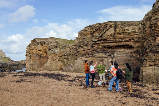 A wide shot of a medium group of young people gathered on a beach at Amble in Northumberland, North East England. They are on a field trip, talking about the rock formations which they are standing beside. They are wearing casual clothing and behind them is a large rocky outcrop, whilst seaweed coats the beach surface.