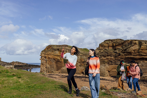 A shot of a group of young people walking along a beach at Amble in Northumberland, North East England. They are on a field trip, some of them carrying notepads and digital devices. They are wearing casual clothing and behind them is a large rocky outcrop. Two females walk together at the front, one pointing ahead of them.
