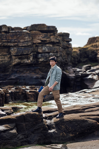 A shot of a young adult male walking across a rocky area at the edge of the beach at Amble in Northumberland, North East England. He is holding a notebook and looking directly towards the camera, wearing casual outdoors clothing. It is a sunny day.