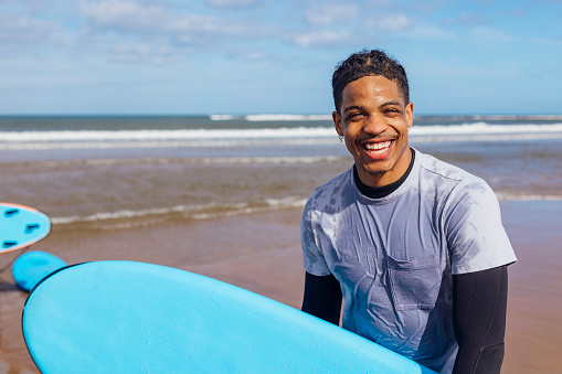 A shot of a young male adult standing on the beach holding a surfboard at Amble in Northumberland, North East England. He is smiling, wearing a wetsuit with rash vest over. Behind him is a large expanse of sand and it is a sunny day.