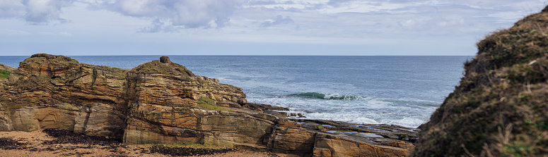 A panoramic shot of a rugged coastline at Amble in Northumberland, North East England. There are several rocky outcrops, behind which there are tidal pools and the sea beyond. There is a wave rolling in towards the rocks and the sea is calm.