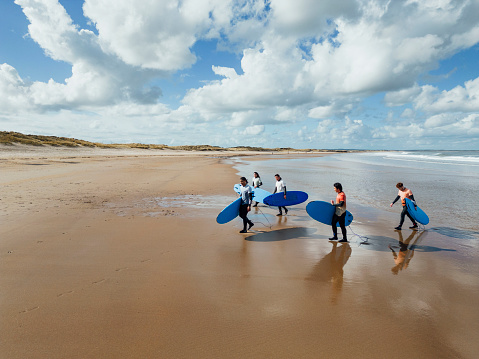 A wide shot of a group of people walking with surfboards on the beach at Amble in Northumberland, North East England. They are walking in from the sea after a surfing session with their instructor, wearing wetsuits and rash vests. It is a sunny day.