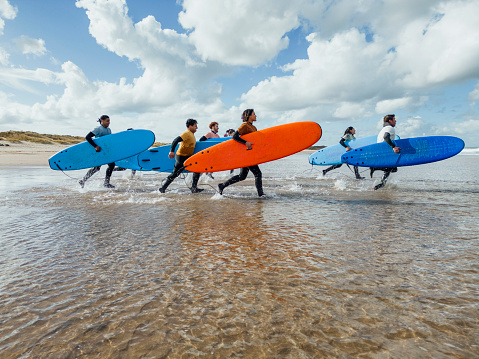 A shot of a medium group of people walking with surfboards on the beach at Amble in Northumberland, North East England. They are walking in from the sea after a surfing session with their instructor, wearing wetsuits and rash vests. It is a sunny day.