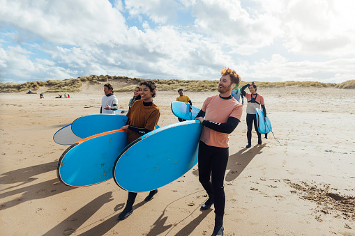 A shot of a group of people walking with surfboards on a beach at Amble in Northumberland, North East England. They are walking towards the sea ready for a surfing session with their instructor, wearing wetsuits and rash vests. It is a sunny day.