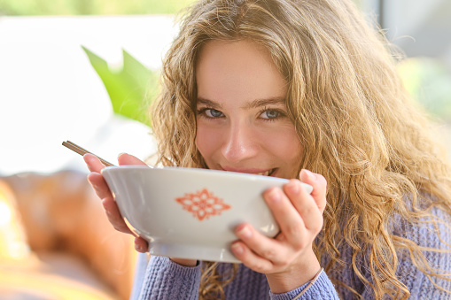 Young glad blond woman eating ramen from bowl and looking at camera on blurred backdrop