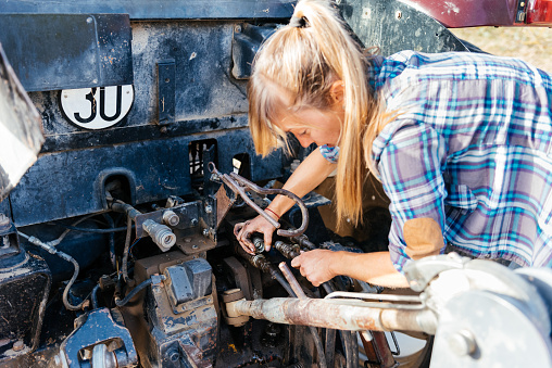 Side view of female mechanic in casual clothes fixing cables on tractor while working in agricultural field during summer day