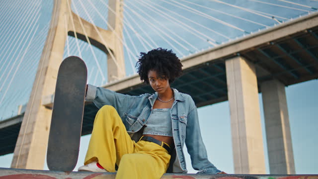 Bright girl posing skateboard at urban bridge. African skater sitting street