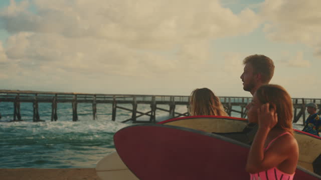 SLO MO Surfer friends walking with surfboards on ocean beach along pier