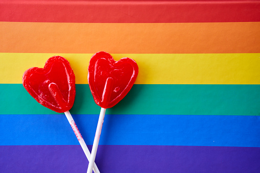 Two red heart-shaped lollipops with a background of the lgbtiq+ flag, representing love.