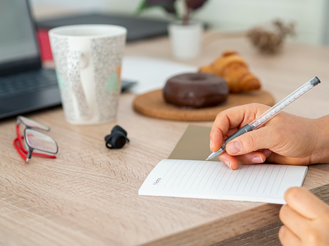 Close-up of woman hand holding pen with a notebook ready to write and planning a to-do list. Working desk with a coffee cup, snack and glasses during a brainstorming session. Taking notes
