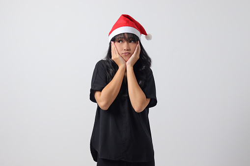 Portrait of shocked Asian woman with red Santa hat holding hands on cheeks, showing surprised and scared expression. New year and christmas concept. Isolated image on white background