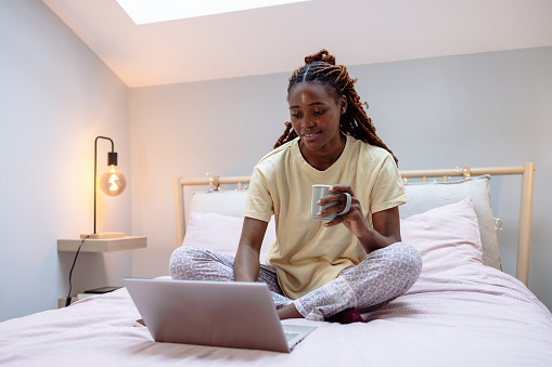 A young African American woman relaxing in her bedroom in a lotus position, while holding a coffee mug in her hand. She is in her pajamas with a soft smile on her face.