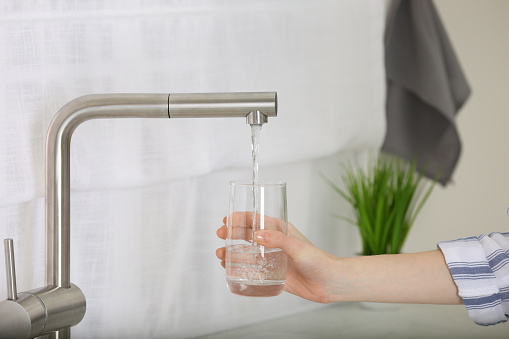 Woman filling glass with tap water from faucet in kitchen, closeup