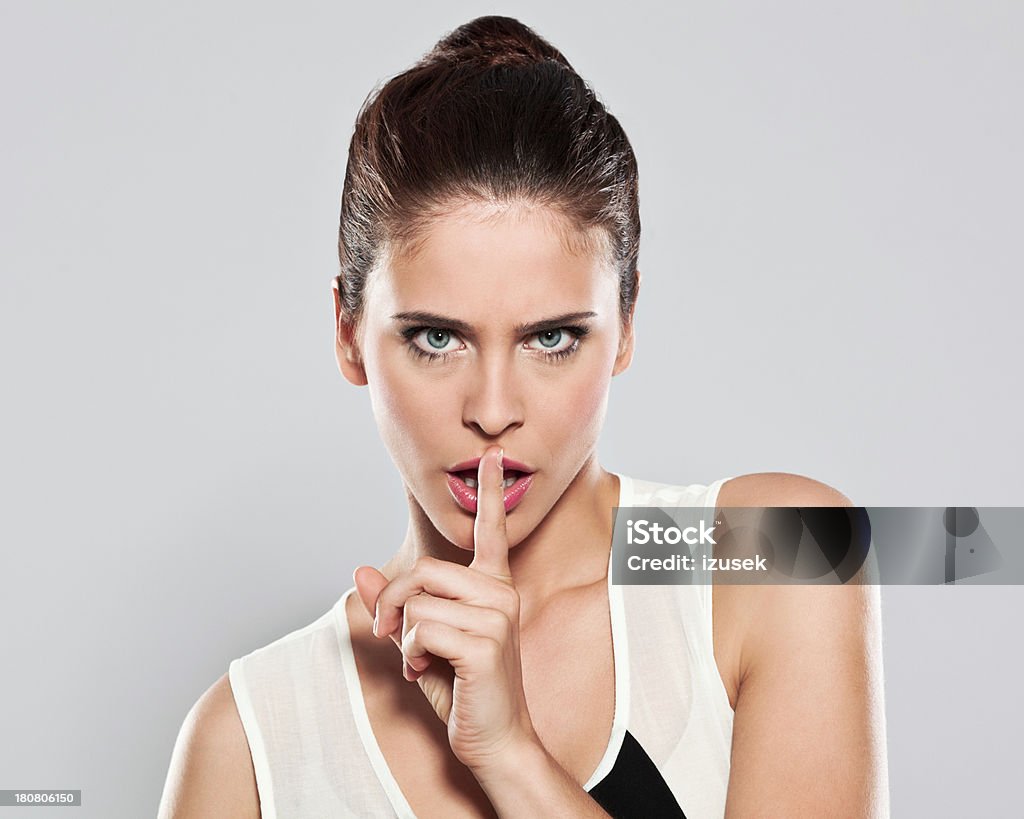 Attractive young woman shushing Portrait of beautiful young woman looking at camera with finger on lips. Studio shot on a grey background. 20-24 Years Stock Photo