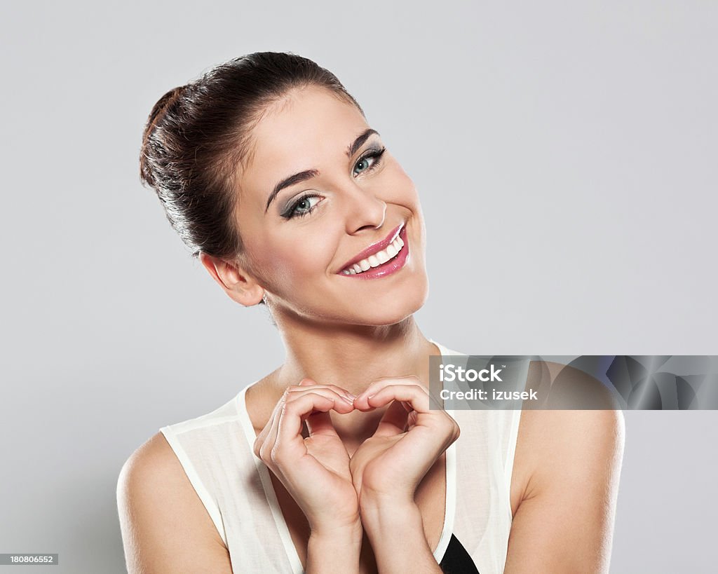 Happy young woman, Studio Portrait Portrait of cheerful young woman making heart shape with hands and smiling at the camera. Studio shot on a grey background. Dental Health Stock Photo