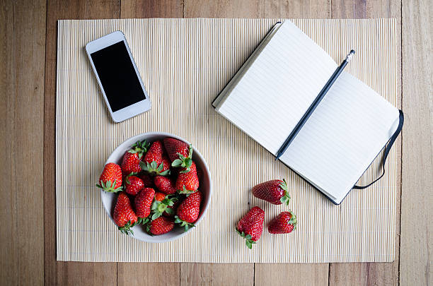 Blank recipe book, modern smartphone and a bowl of strawberries on a wood table.