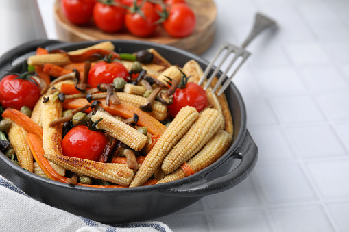 Tasty roasted baby corn with tomatoes, capers and mushrooms on white tiled table, closeup