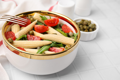 Tasty baby corn with tomatoes, arugula and capers on white tiled table, closeup