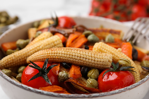 Tasty roasted baby corn with tomatoes and capers on table, closeup