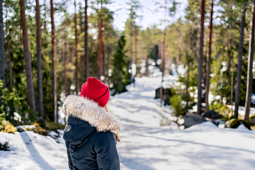Winter snow in forest. Woman in nature outdoors. Red hat and warm coat with fur hood. Behind back portrait of girl. Cold sunny day. Morning sunlight in Finland. Walk path outside  in the woods.