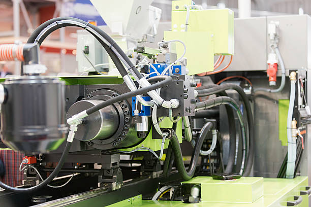 A industrial plastic injection molding machine.  The machine features gray tubes, clear plastic joiners, red wires and various other parts.  The close-up view of this industrial machine creates an abstract effect with a mixture of gray, green, red, blue and yellow colors.