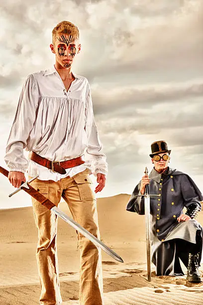 Two rag tag soldiers ready for battle should it present itself.  Swords in hand anxious for the fight to come to them.  Sand Dunes behind and in the forefront.  Cloudy omnious skies.  One man forward and one man behind waiting for instructions.