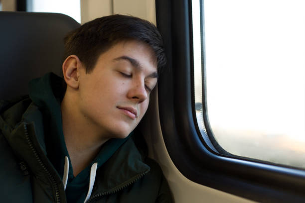 Young brunette man in a warm jacket sits by the window on a train and sleeps peacefully, leaning against the wall. Traveler in a train with his eyes closed sits tired on the road