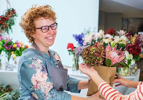 Positive female florist in glasses and apron giving bouquet of fresh blooming hydrangea and protea in paper bag to customer during workday in floral shop