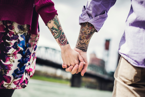 A closeup of a couple holding hands along the Willamette river waterfront in Portland, Oregon, looking at the view of the city.  Horizontal, with an emphasis on their forearm sleeve tattoos.