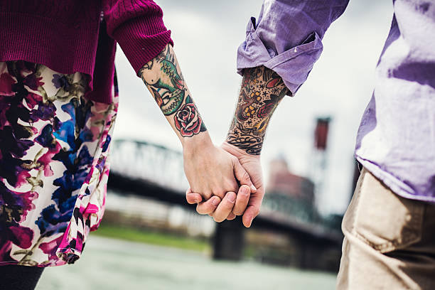 Modern Couple Holding Hands on Portland Waterfront A closeup of a couple holding hands along the Willamette river waterfront in Portland, Oregon, looking at the view of the city.  Horizontal, with an emphasis on their forearm sleeve tattoos. body adornment rear view young men men stock pictures, royalty-free photos & images