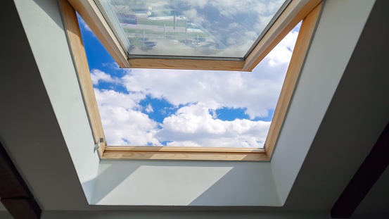 Looking on clouds flying past at the blue sky through open attic window in house roof.
