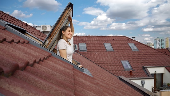 Happy smiling brunette woman opening attic window and breathing fresh air while looking on the cityscape with red tiled roofs.