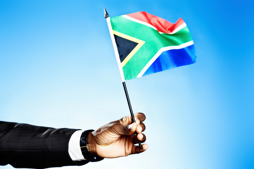 A dark-skinned masculine hand holds up the distinctive South African national flag against a sky-blue background. He is obviously a patriotic citizen!