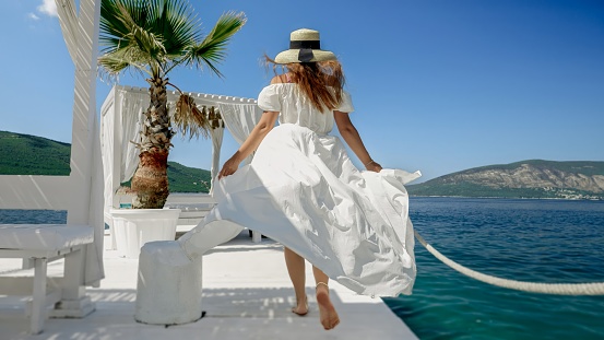 Lady in a flowing light dress and straw hat enjoys a carefree walk on a wooden pier at the beach, evoking the spirit of a summer vacation.