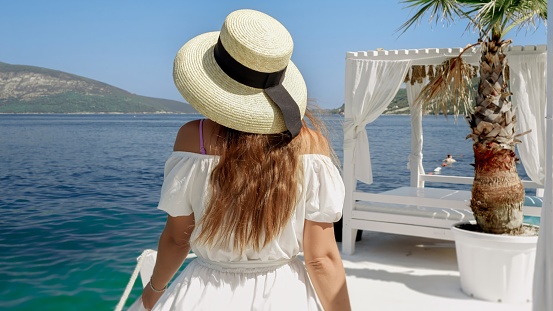 Rear view of elegant woman in hat and long dress relaxing by walking at the wooden sea pier on a sunny summer day.