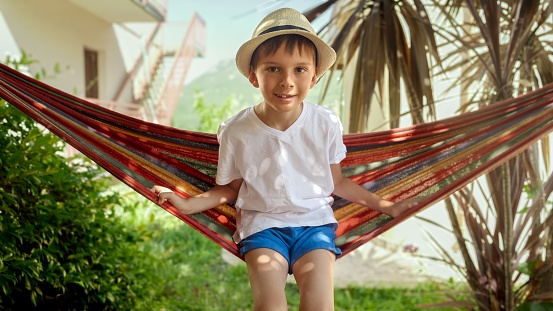 Portrait of little boy sitting in hammock and swaying at garden. Carefree childhood memories, and the relaxation of a vacation.