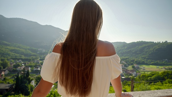 Rear view shot of brunette woman enjoying view on alley and mountains with sunset light from her villa terrace.