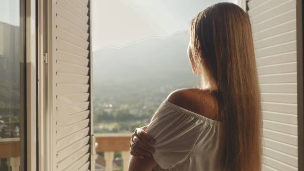 istock Young brunette, dressed elegantly, opens her window blinds, ventures to the terrace, and looks at the mountain sunrise or sunset a serene stock footage capturing vacation bliss 1808199542