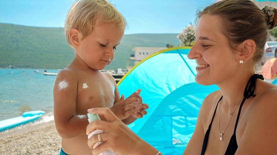 Young mother carefully applies sunscreen spray to her baby son's delicate skin as they relax on a sunny sea beach. Importance of healthcare during summer vacations with kids.