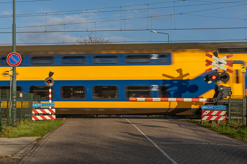 Bunnik, the Netherlands 12 November 2023. Railway crossing on a rural road, stop sign and warning lights, crossing barriers and a train passing, farms and Dutch countryside in background, sunny autumn