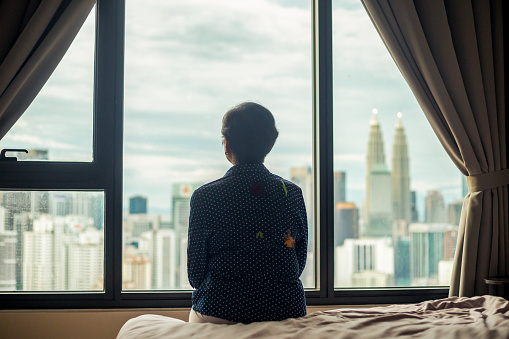 Silhouette of senior woman looking through window from apartment with city view