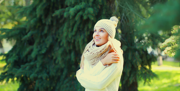 Winter portrait of happy smiling blonde young woman wearing hat, knitted sweater and scarf in forest on christmas tree background