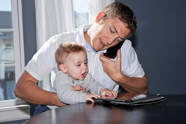 Father multi-tasking with baby Father (20s) on mobile phone, using digital tablet with baby (8 months) on lap. Sc0601 stock pictures, royalty-free photos & images