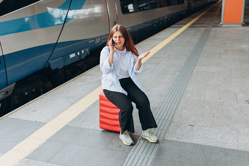 Young angry redhead woman waiting train on station platform, she lost or late, unhappy girl talking on smart phone. Railroad transport concept