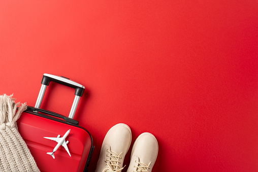 Embrace the New Year spirit with a picturesque setting: top view of white winter boots, a suitcase, plane model, and a knitted scarf on a vibrant red backdropâperfect for New Year travel promos